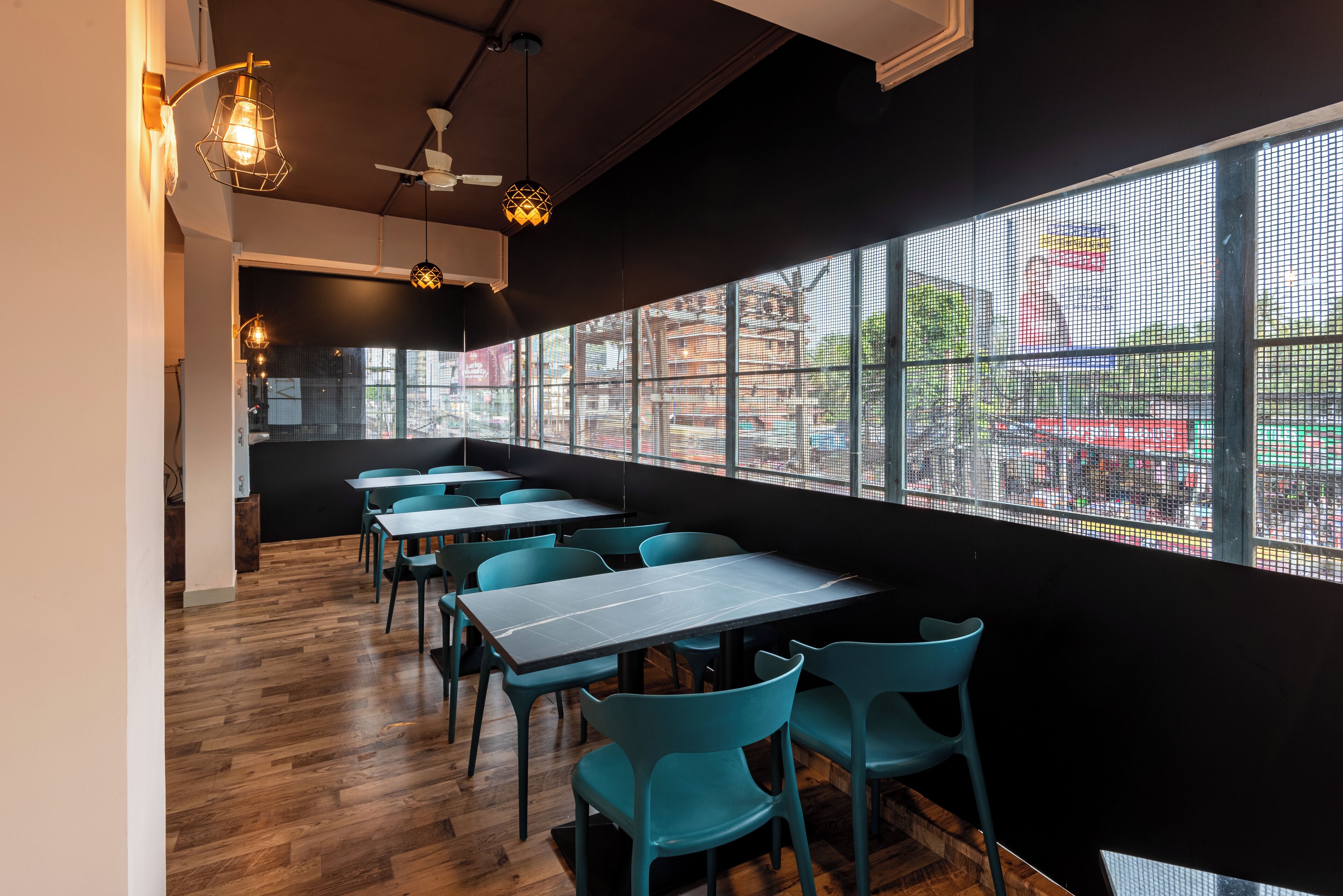 A modern restaurant interior with wooden flooring, teal chairs, and tables, featuring large windows with a city view.