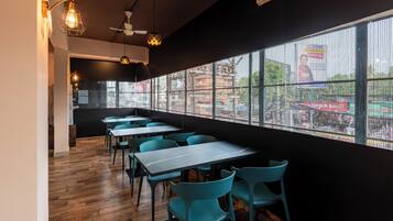 A modern restaurant interior with wooden flooring, teal chairs, and tables, featuring large windows with a city view.