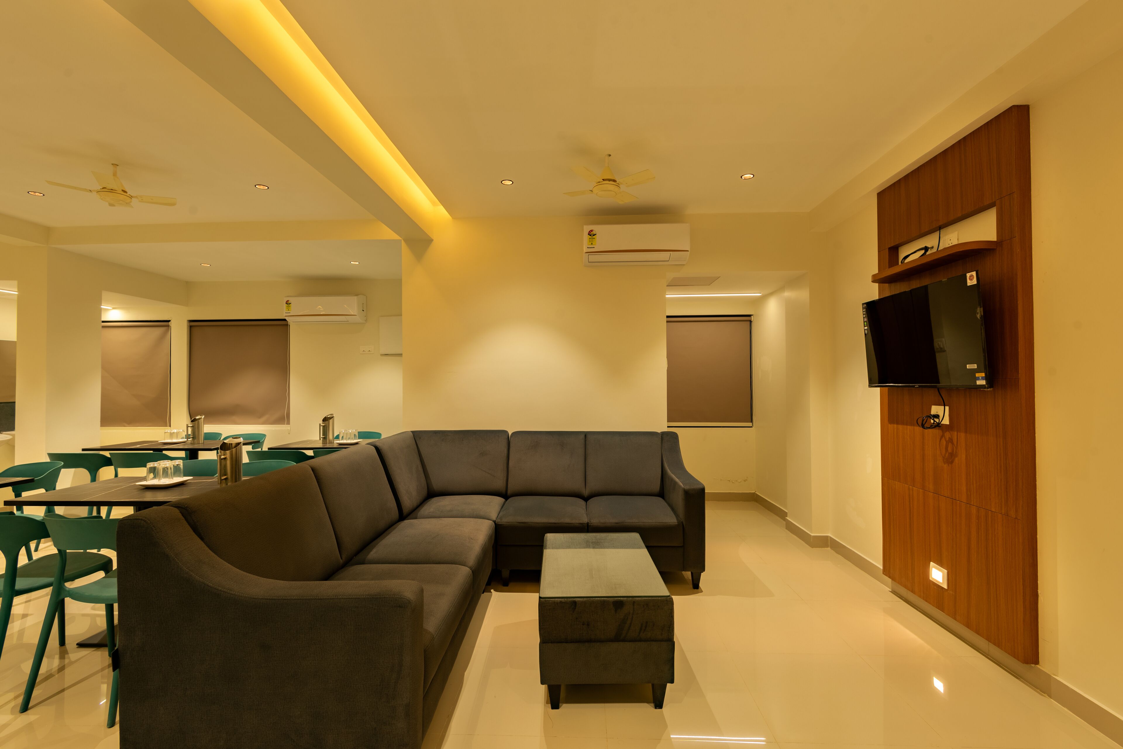 A modern living room with a sectional sofa, a coffee table, and a flat-screen TV mounted on a wooden panel. There are ceiling fans, recessed lighting, and air conditioning units.