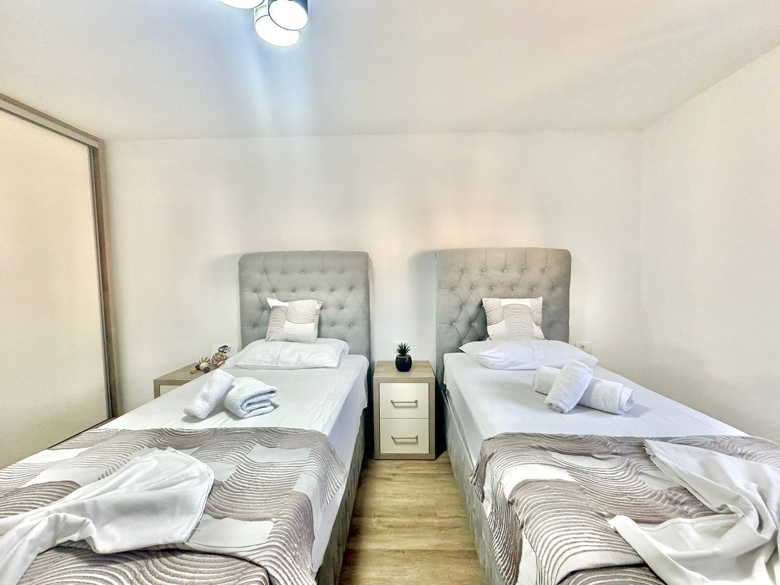 Et hotelværelse med to senge, hver med en betrukken sengegavl, hvide sengetøj og stribede plaider. Et natbord placeret mellem sengene har en lille potteplante.
