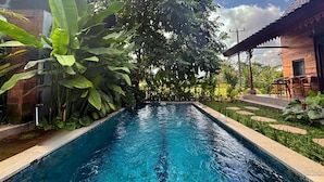 A rectangular swimming pool with clear blue water, surrounded by lush green plants and trees.