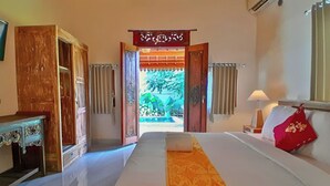A hotel room with a bed, a wooden wardrobe, a desk, and a view of a garden through an open doorway.