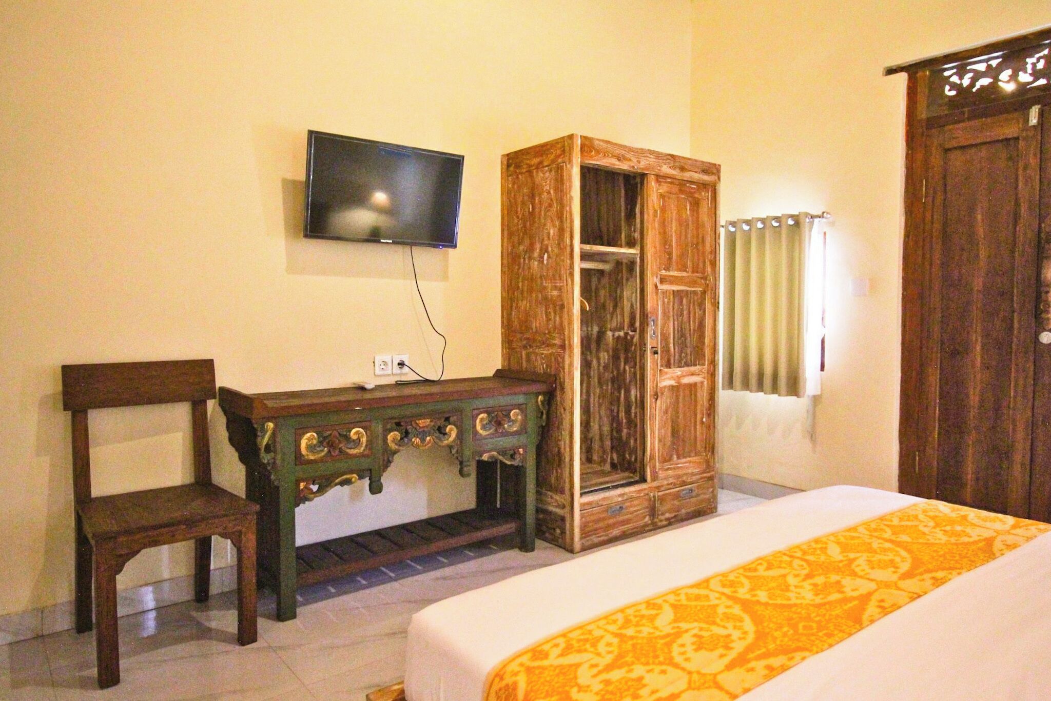 A bedroom with a bed, a wooden wardrobe, a television mounted on the wall, and a desk with a chair.