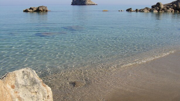 A clear coastal beach with calm waters, rocky outcrops, and a sandy shore.