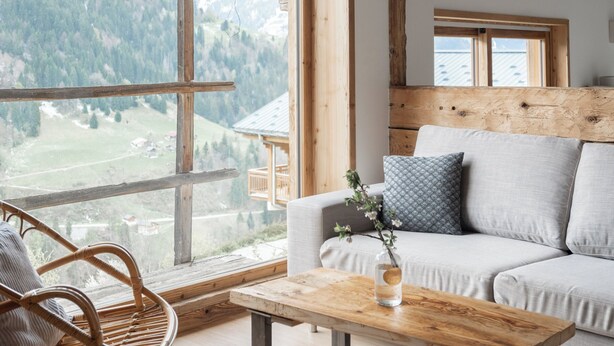 Ein gemütliches Zimmer mit Holzbalkendecke, einem großen Fenster mit Bergblick, einer Couch mit Kissen und Korbsesseln.