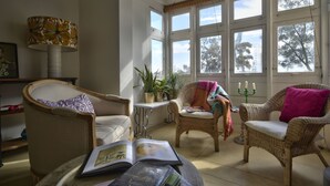 A cozy living room with wicker furniture, a table with books, and large windows with a view of trees. - Sea Breeze Apartment (Floriana)