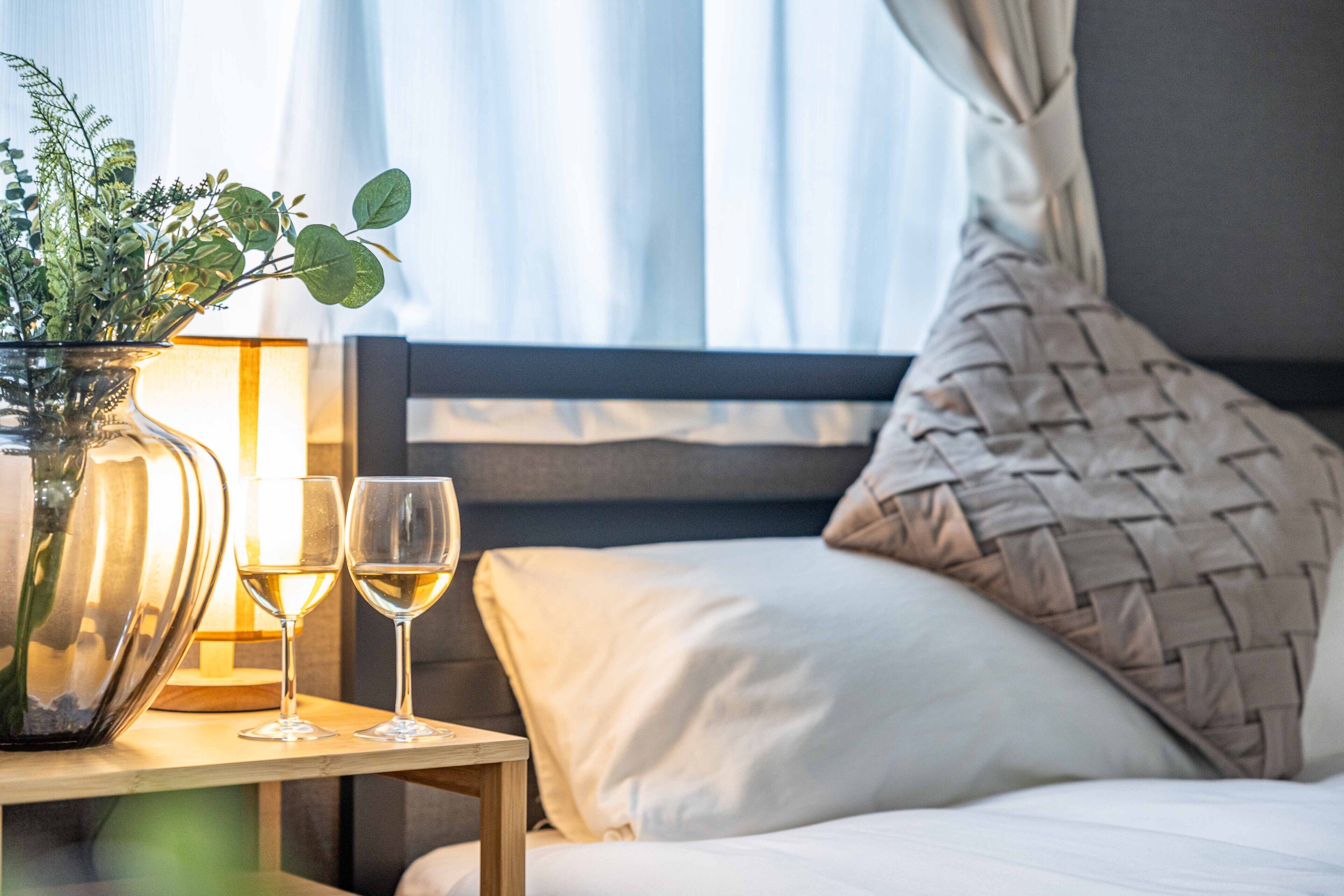 Un lit bien fait, avec un coussin en tissu tissé, une table de chevet ornée d’un vase rempli de verdure et de deux verres de vin blanc, et une lampe.
