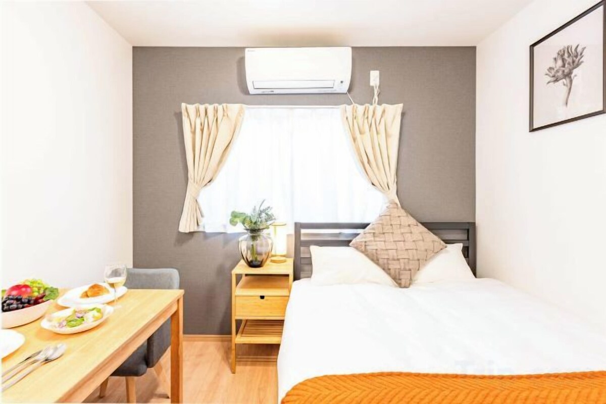 A hotel room with a bed, a bedside table with a lamp, a window with curtains, a small table with a bowl of fruit, and a framed picture on the wall.