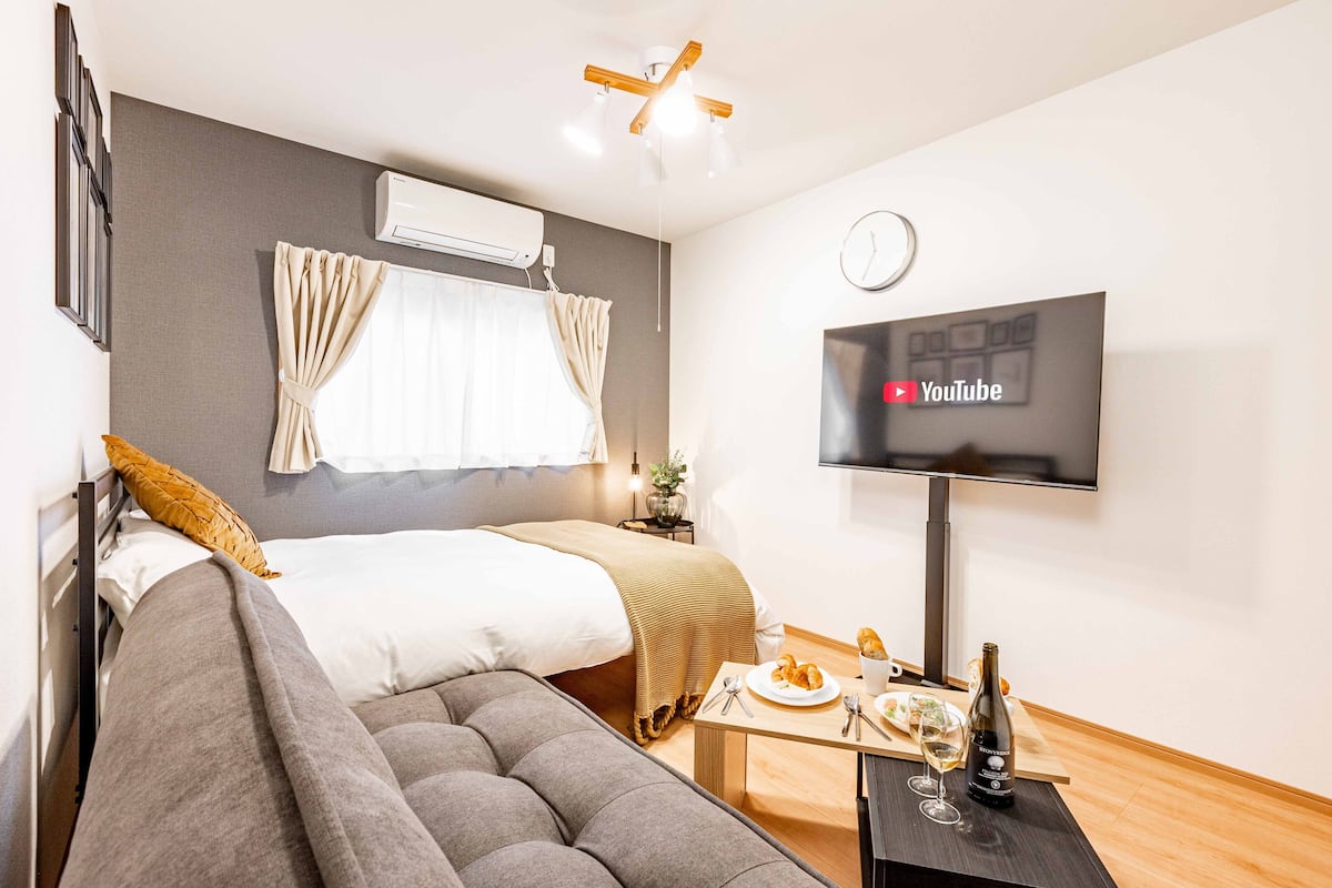 Quarto de hotel moderno com cama, sofá, televisão e uma mesinha com garrafa e copos.