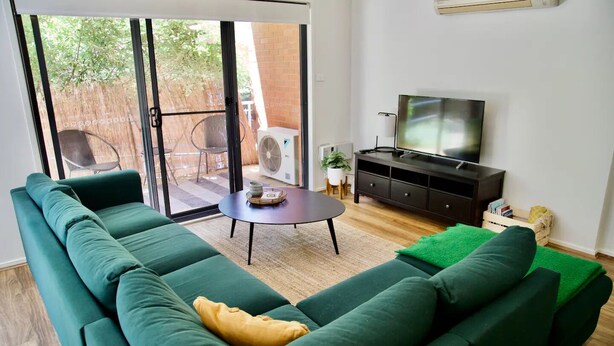Una sala de estar con un sofá verde, una mesita redonda, un televisor y un ventanal corredero que da a un espacio exterior.