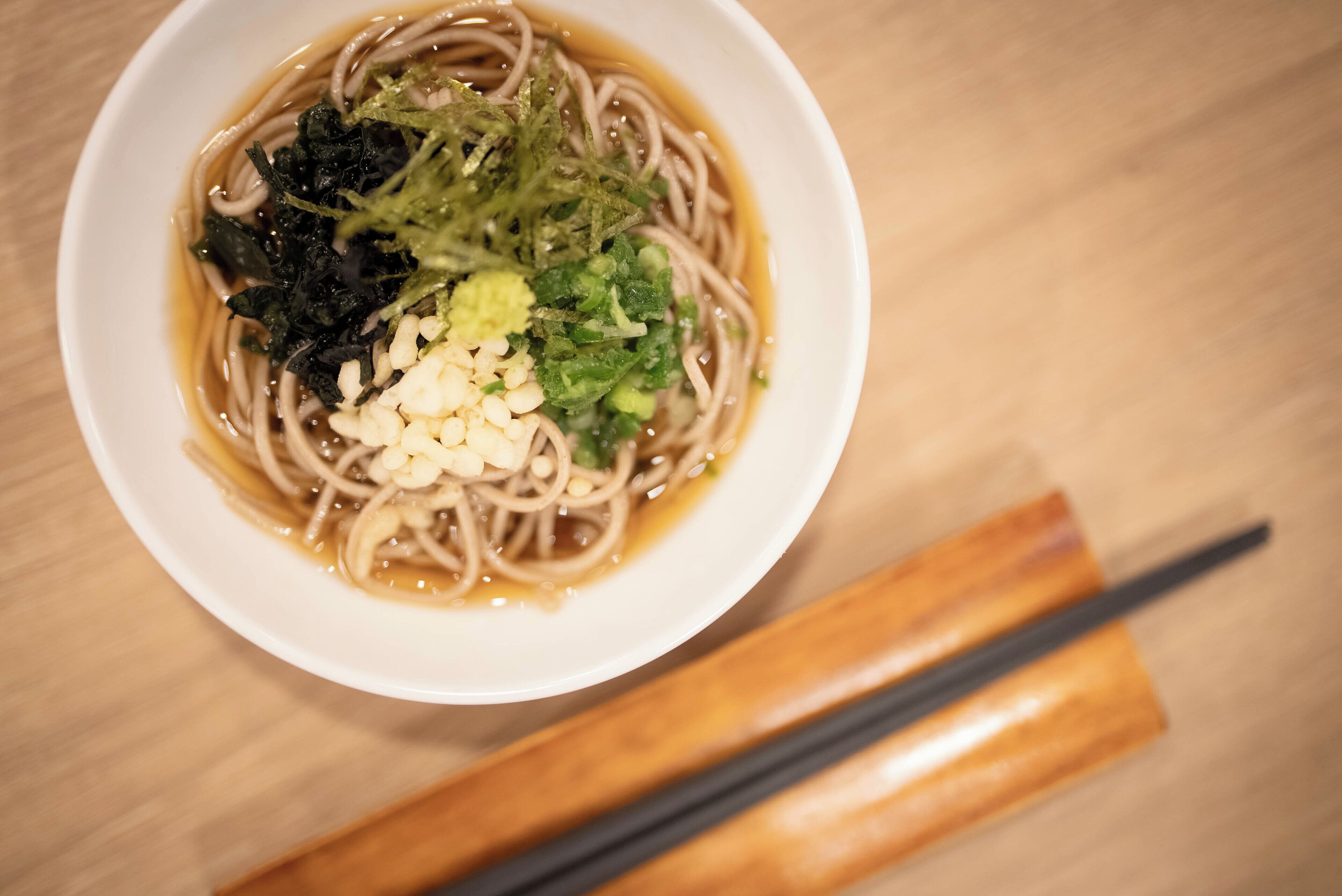 Una ciotola di noodles con condimenti e bacchette su un tavolo di legno.