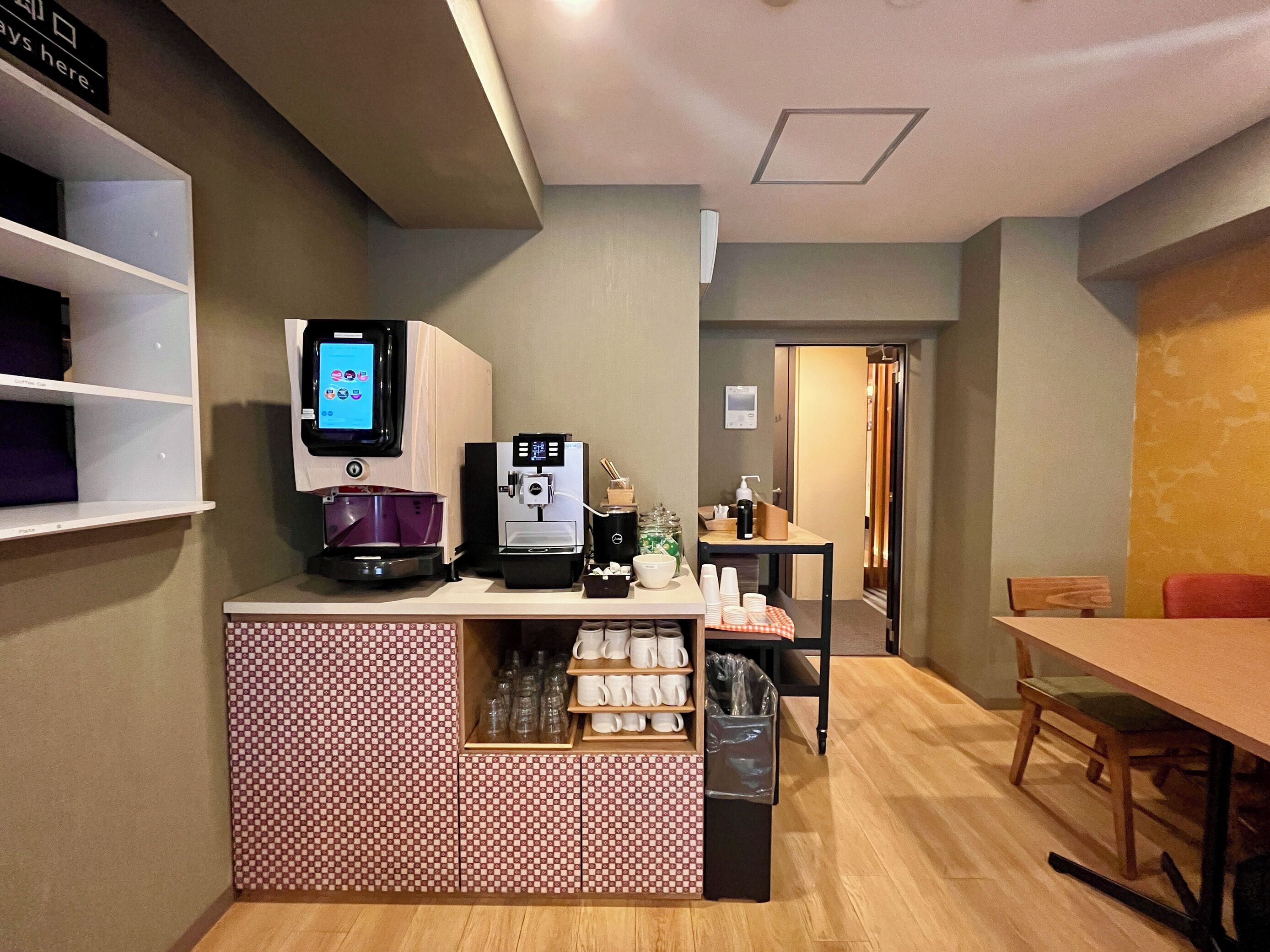 A small coffee station with two coffee machines, a digital screen, and a counter with cups and a trash bin.