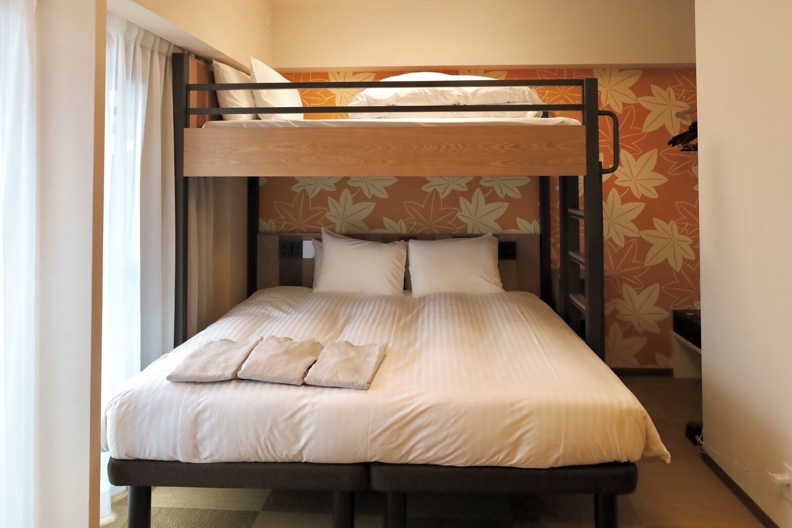 A bunk bed with a wooden frame and white bedding, a window with curtains, and a wall with a floral pattern.
