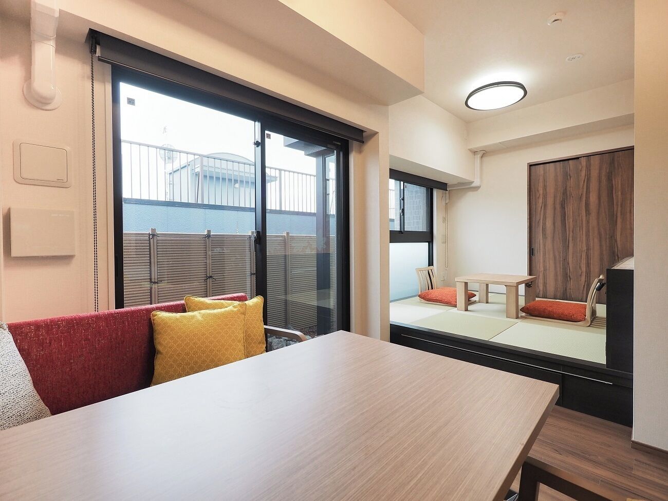 A modern room with a wooden table, red and yellow cushions, and a sliding glass door leading to a balcony with a view of a fence and buildings.