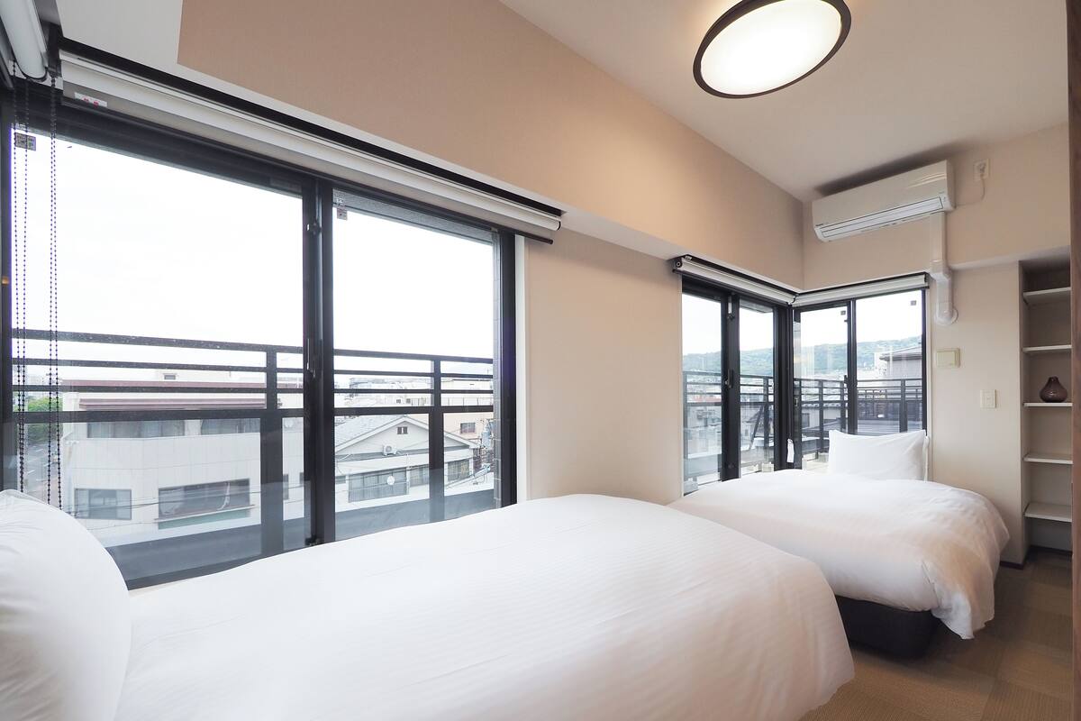 A hotel room with two beds, large windows, a ceiling light, and a view of the city.