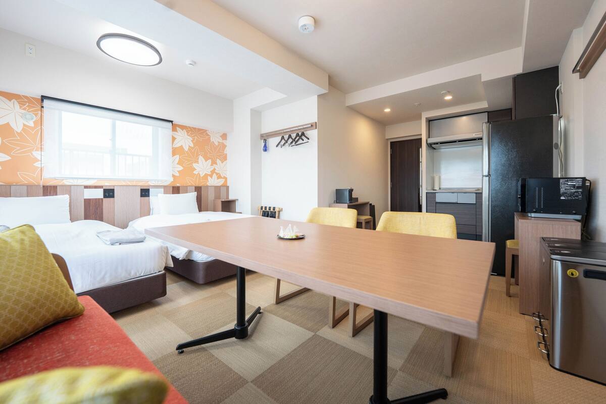 A hotel room with a bed, a sofa, a dining table, a kitchen area with a refrigerator, and a window with a flower pattern.