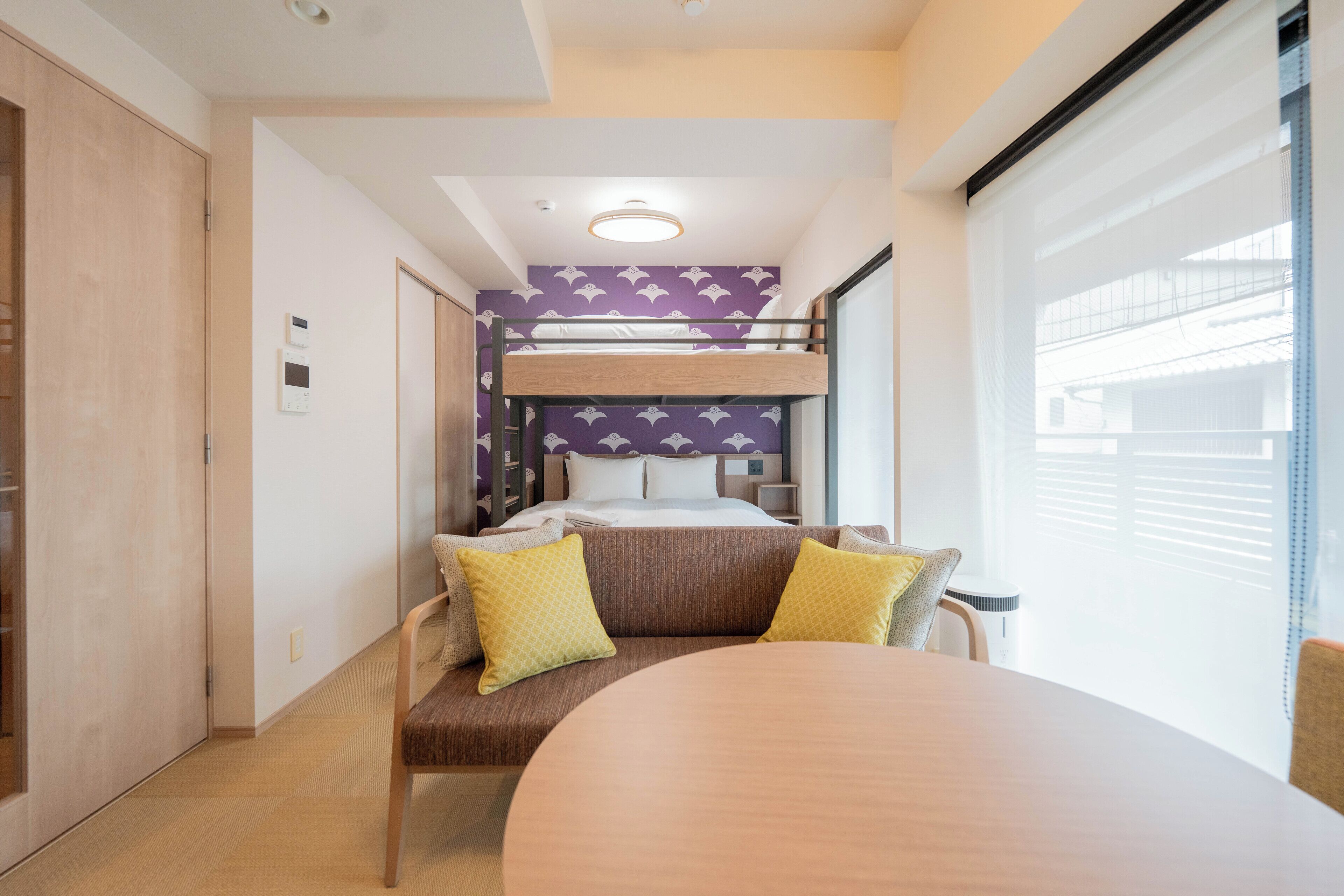 A compact hotel room with a bunk bed, a round table, and a sofa with yellow cushions.