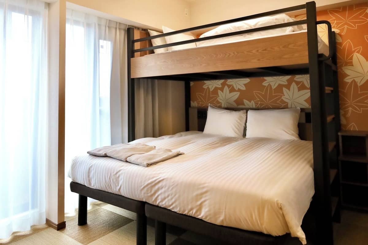 A bunk bed with a wooden frame and white bedding, a window with sheer curtains, and a wall with a leaf pattern.