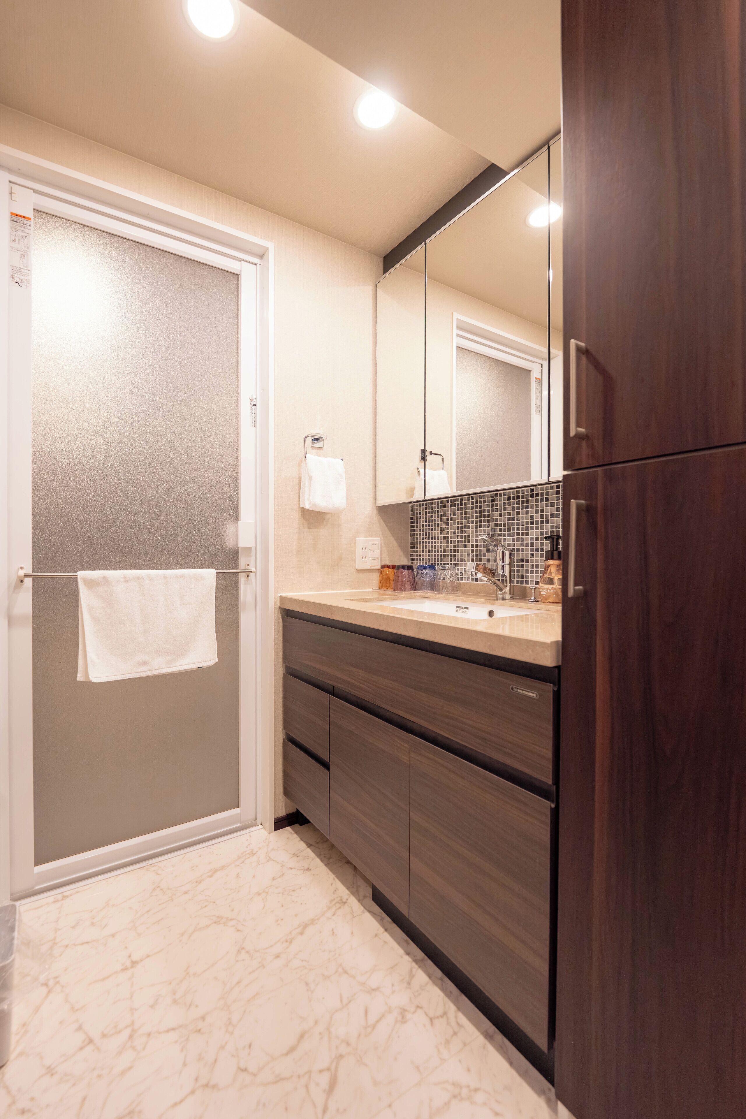 A modern bathroom with a marble floor, a wooden vanity, a large mirror, and a towel hanging on a door.