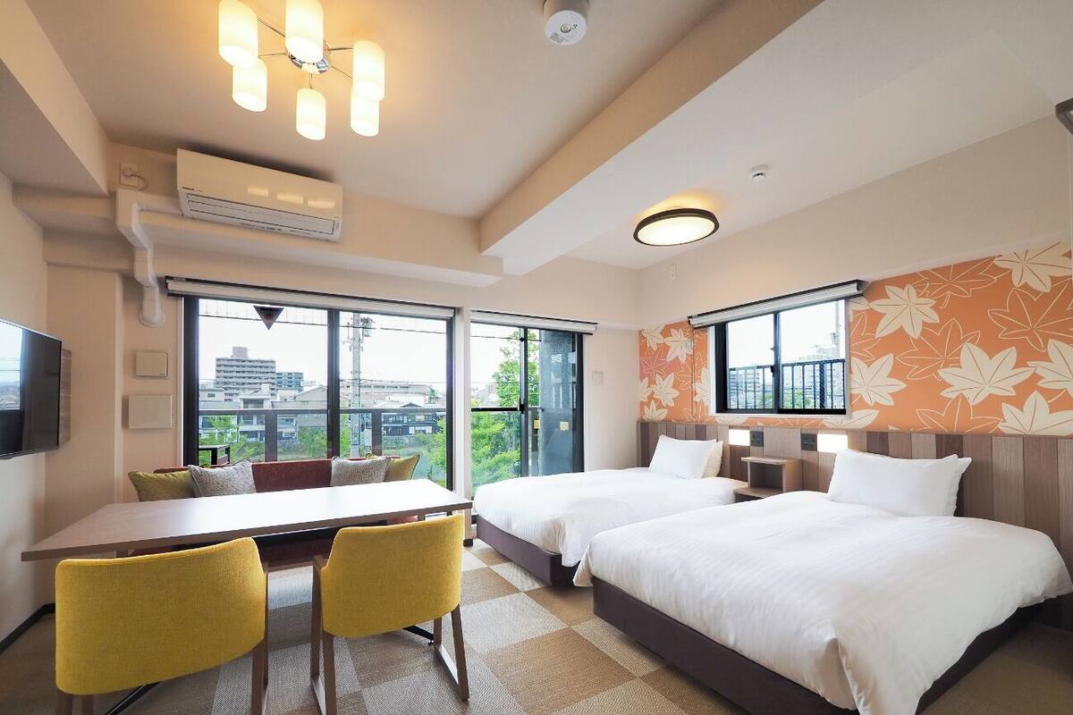 A hotel room with two beds, a dining table with chairs, a TV, and large windows with a city view.