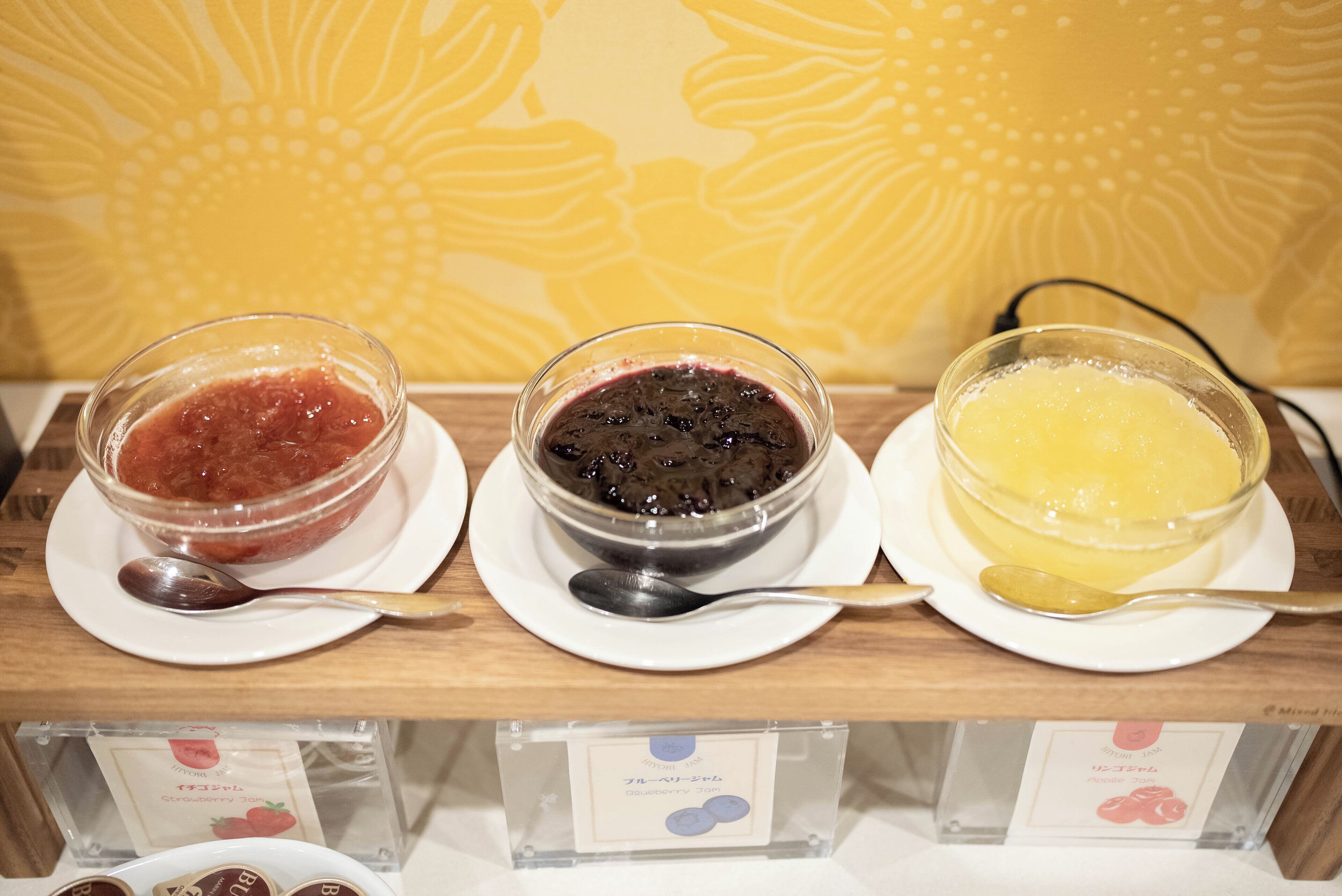 Drei Schalen mit unterschiedlich farbigen Marmeladen auf weißen Tellern, mit Löffeln, vor einem gelben Hintergrund mit Muster.