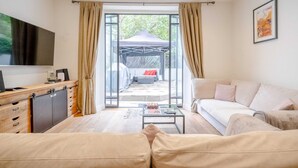 A modern living room with a sofa, a coffee table, and a TV. There is a sliding glass door leading to an outdoor area with a tent and a couch. - Gorgeous 2BD Flat With Patio - South Kensington! (London)