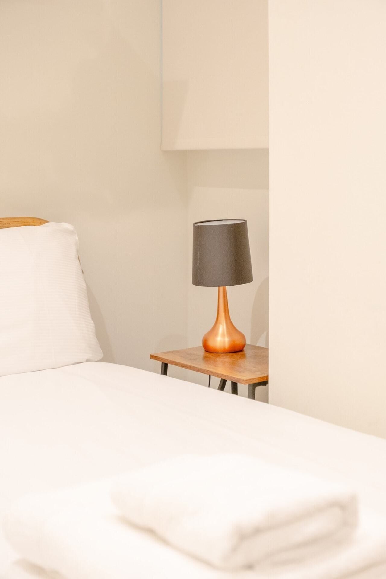 A neatly made bed with a white comforter and pillows, a small wooden nightstand with a lamp, and a white wall in the background.