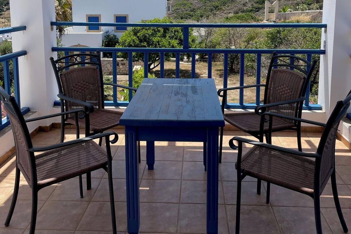 A covered outdoor seating area with a table and four chairs, overlooking a hillside with a white building.