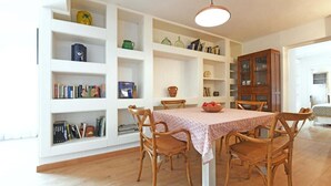 Zona pranzo con tavolo ricoperto da una tovaglia fantasia, circondato da sedie in legno. Si vedono scaffali bianchi con diversi oggetti, tra cui libri e decorazioni. È visibile anche un mobile in legno con vetrine.