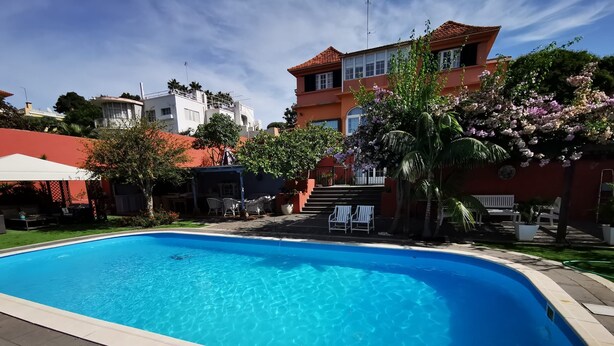 Una piscina con bordo curvo, circondata da una terrazza con mobili da esterno e una scala che conduce a un edificio con tetto rosso.