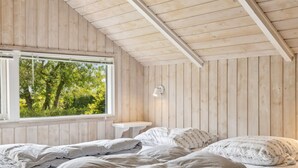 Une chambre avec un grand lit, une petite table de chevet, une fenêtre donnant sur des arbres et des murs en bois.