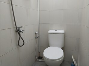 Une petite salle de bain aux murs recouverts de carreaux blancs, équipée de toilettes, d’un flexible de douche et d’un robinet mural.