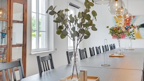 Uma longa mesa de jantar com cadeiras, vasos de vidro transparente com folhagem verde e um tabuleiro de madeira com pequenas xícaras e recipientes para temperos.