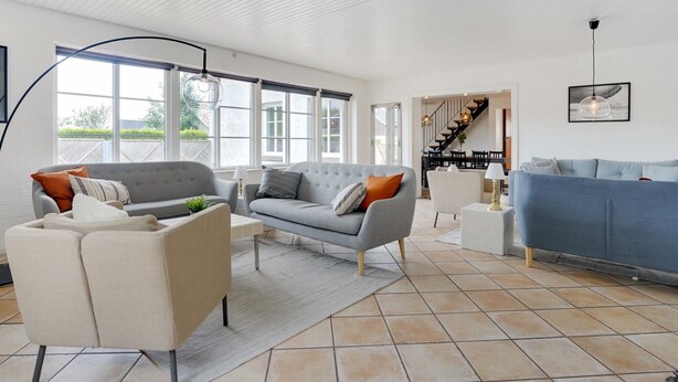 Uma sala de estar espaçosa com um sofá cinza, uma poltrona bege e outro sofá azul. Há uma escada que leva a um andar superior e grandes janelas que proporcionam luz natural.