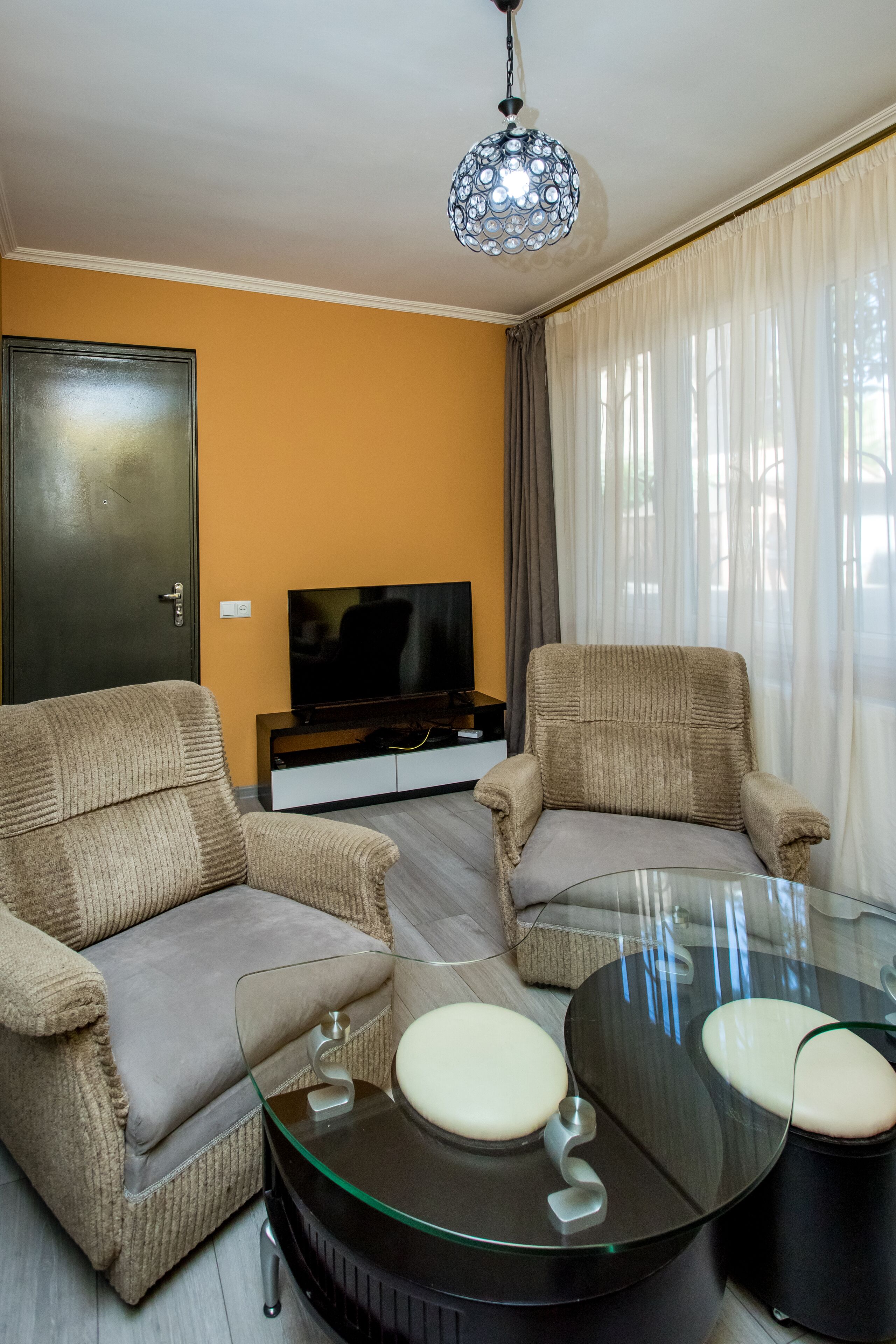Uma sala de estar com dois poltronas bege, uma mesa de centro de vidro, uma televisão e uma porta.