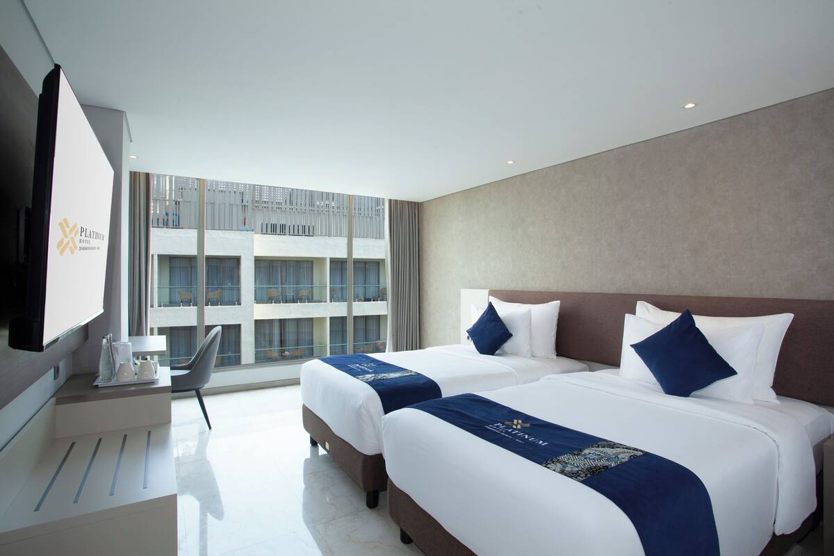 A hotel room with two beds, a flat-screen TV, a desk with a chair, and a large window offering a view of the building's exterior.