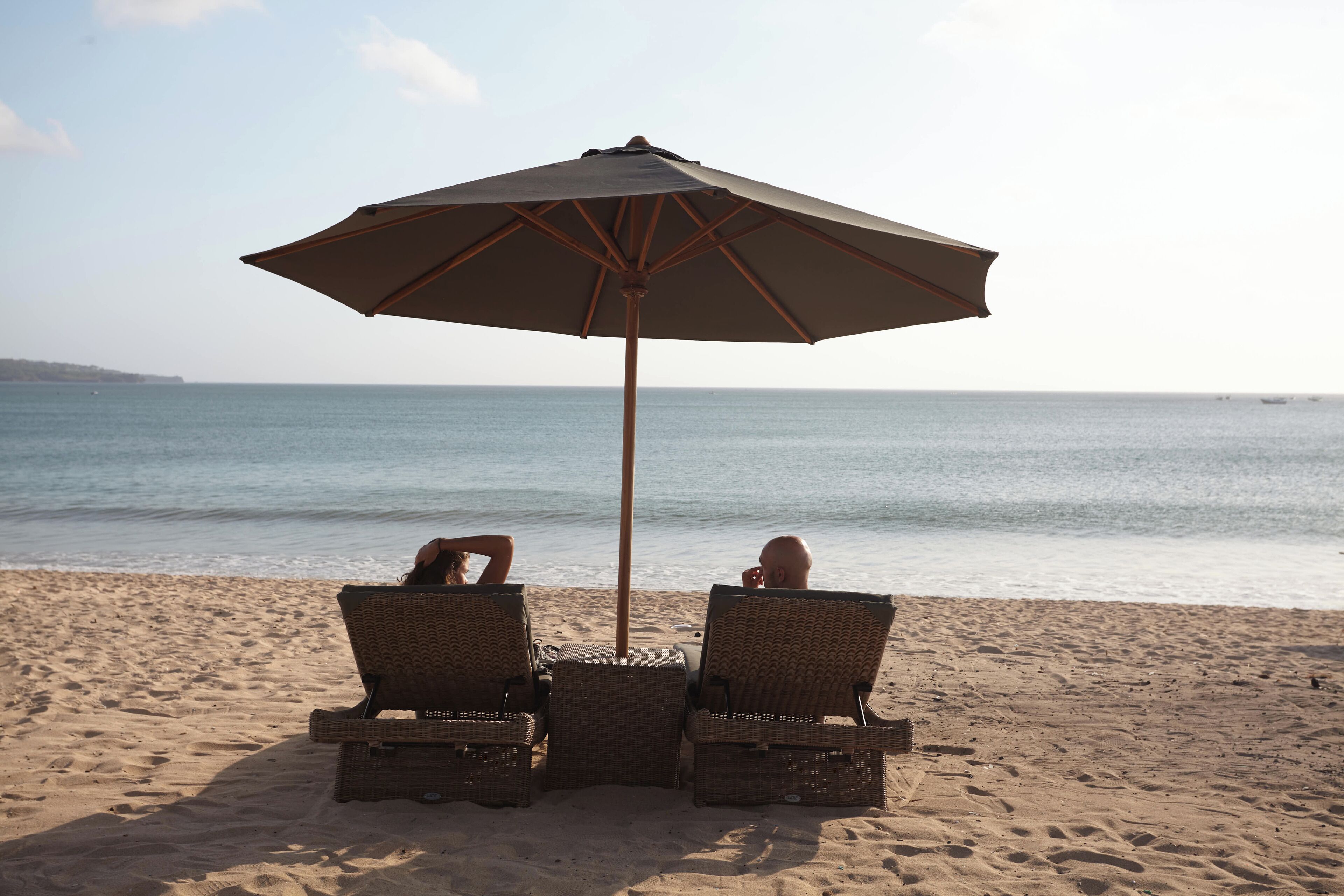 Dos sillones de mimbre bajo un paraguas grande en una playa de arena, con el océano al fondo.