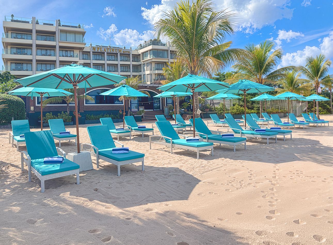 Een strand met rijen ligbedden en parasols, met een hotel op de achtergrond en palmbomen.