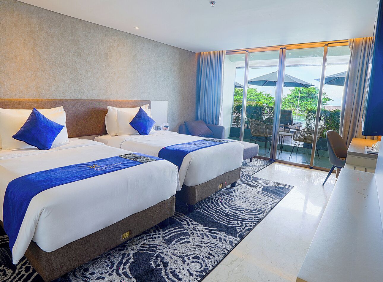 A hotel room with two beds, a large window with a view, a seating area with a table and chairs, and a small desk with a chair.