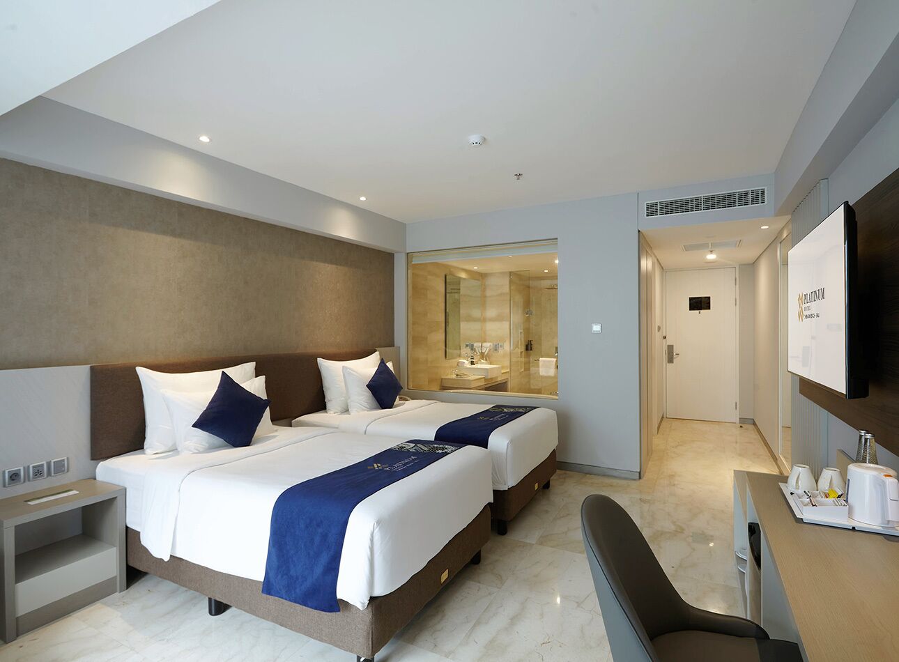 A hotel room with two beds, a desk, a TV, and a bathroom visible through a glass door.