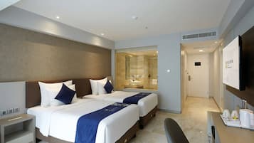A hotel room with two beds, a desk, a TV, and a bathroom visible through a glass door.