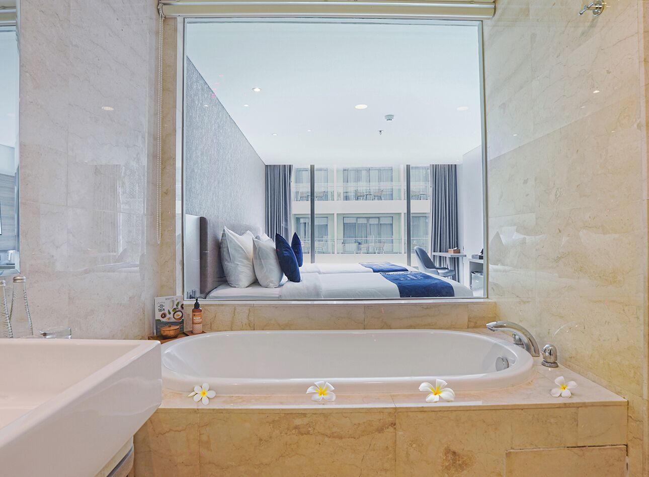 Une salle de bain moderne dotée d’une grande baignoire, d’une douche séparée et d’un grand miroir qui reflète une chambre à coucher avec un lit et des rideaux.