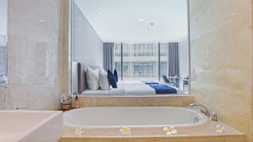 Een moderne badkamer met een ruime badkuip, een aparte douche en een grote spiegel die een slaapkamer met een bed en gordijnen weerspiegelt.
