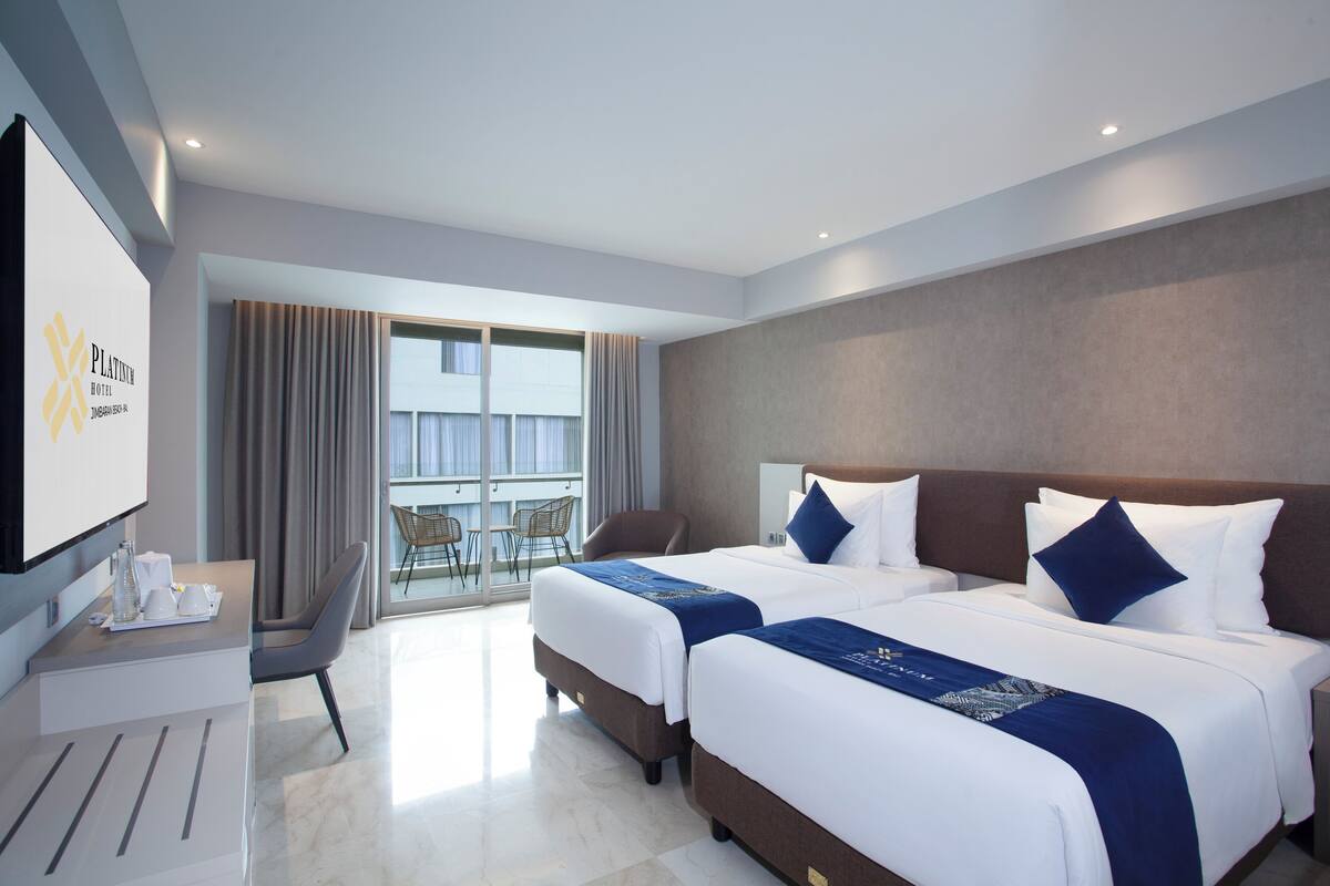 A hotel room with two beds, a desk, a chair, and a large window with curtains.