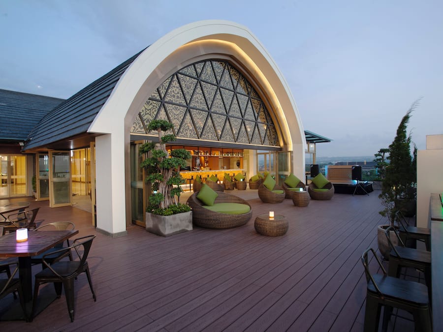 An outdoor seating area with round tables and cushioned chairs, a bar with a large arched window, and a wooden deck.