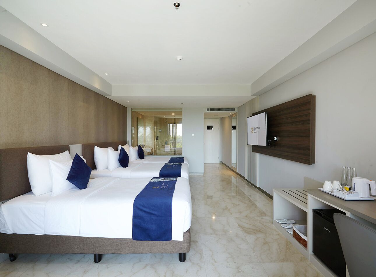 Habitación de hotel con dos camas, un escritorio, un televisor y un baño visible a través de una puerta de cristal.