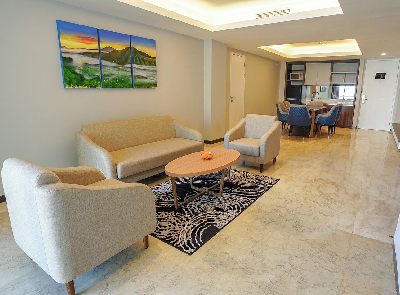 Una sala de estar con un sofá, dos sillones, una mesa de centro y una alfombra. Hay un cuadro en la pared y una zona de cocina al fondo.