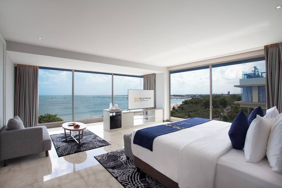 A modern hotel room with a large bed, a seating area, a television, and a view of the ocean.