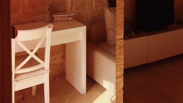 A stone-walled room with a round mirror, a white desk, a chair, and a flat-screen TV on a low wall. - Barakka Garden Studio  (Valletta)