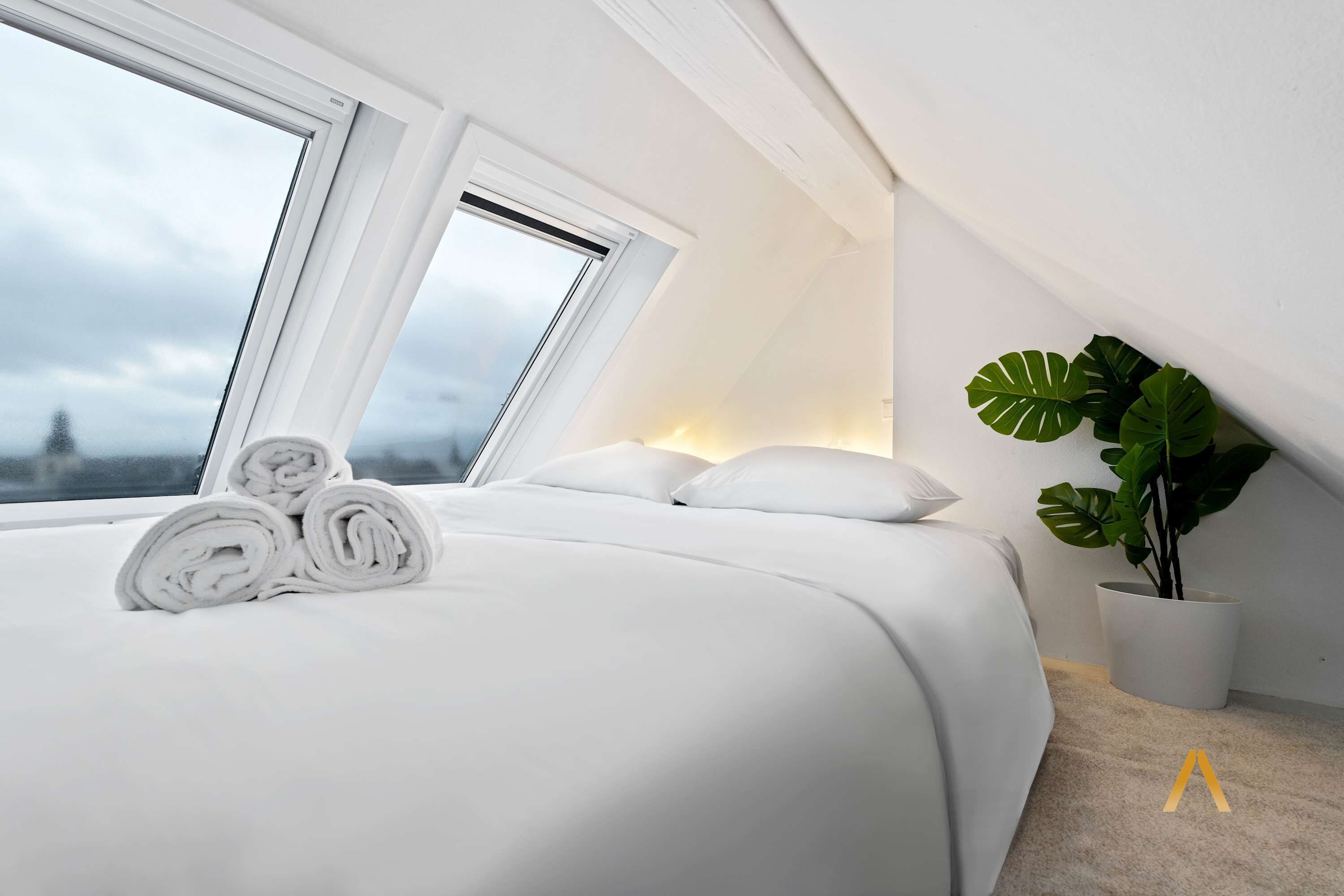 Een net opgemaakt bed met opgerolde handdoeken, een grote kamerplant en uitzicht door een dakraam.